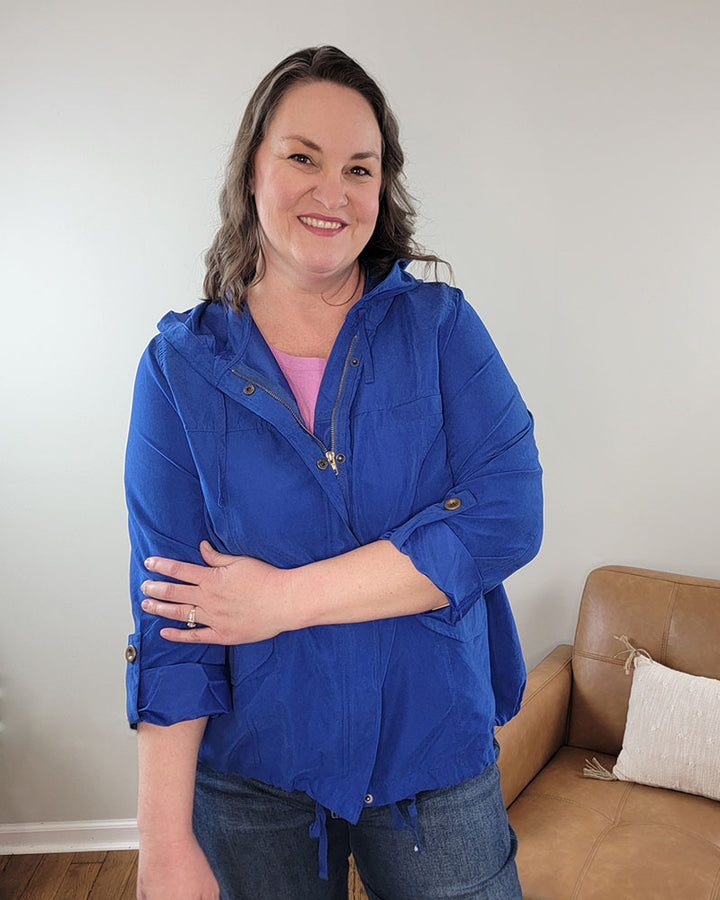 A woman with wavy brown hair smiles, wearing the Alyson Jacket by Mine and E&M in bright blue over a pink top and blue jeans, standing indoors near a tan sofa with a light pillow against a pale wall.