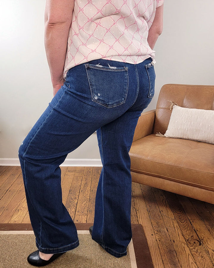 A person wearing Nella Straight Jeans from Risen stands indoors by a tan couch with a white pillow on a wooden floor, facing away from the camera.