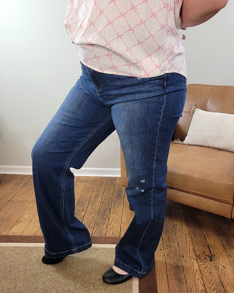 A person is wearing Nella Straight Jeans from Risen, standing indoors on a wooden floor by a tan couch with a white pillow. Only their lower half is visible.