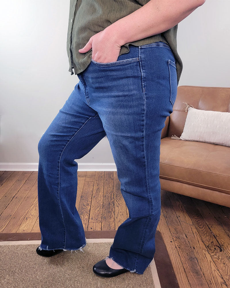 A person wearing Jayda Bootcut Jeans from Lovervet and a green shirt stands indoors with one hand in their pocket, next to a tan sofa with a white pillow on a wooden floor.