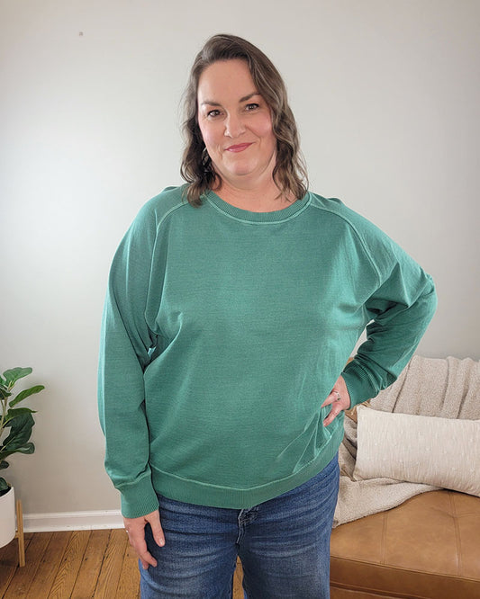 A woman with wavy brown hair smiles indoors, wearing the ZENANA Nelly Pullover Crewneck—a loose, dark green 100% cotton sweatshirt—and blue jeans. She stands by a tan couch with a white pillow and a small potted plant nearby.