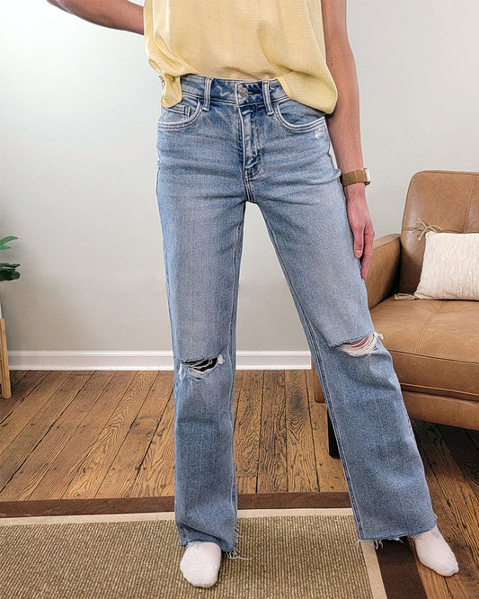 A person wearing Lovervet’s Glorietta Slim Wide Leg Jeans in a light wash and a yellow top stands indoors on a wooden floor beside a tan armchair, with only their torso and legs visible.