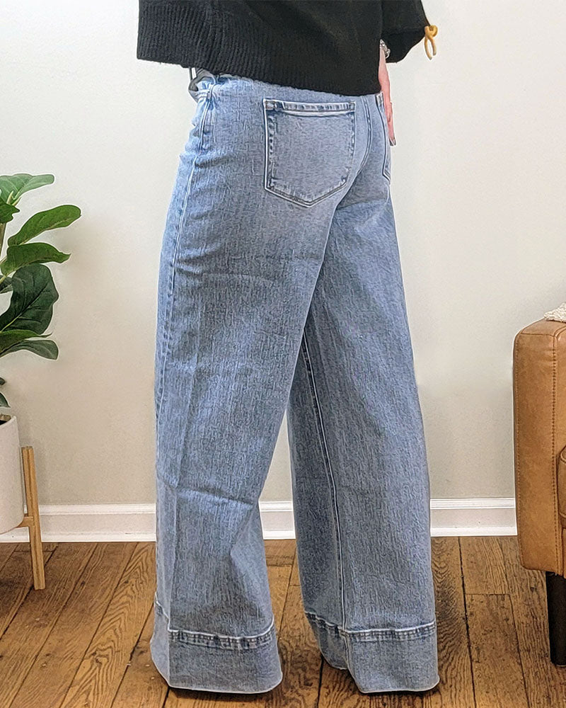 A person wearing Vervet's Aprils Daydream Wide Leg Jeans stands on a wooden floor indoors, next to a green potted plant and a tan sofa. Only their lower half is visible.