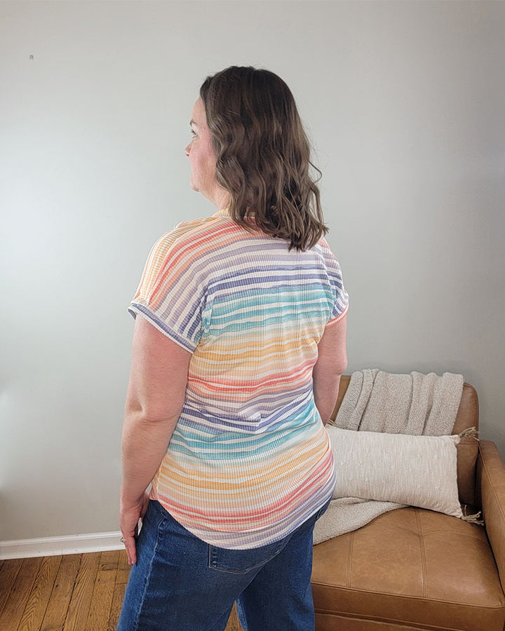 A woman with wavy brown hair faces away indoors, wearing the Heimish Hilary Striped V-Neck tee with pastel rainbow stripes and jeans near a tan couch draped in a light blanket.