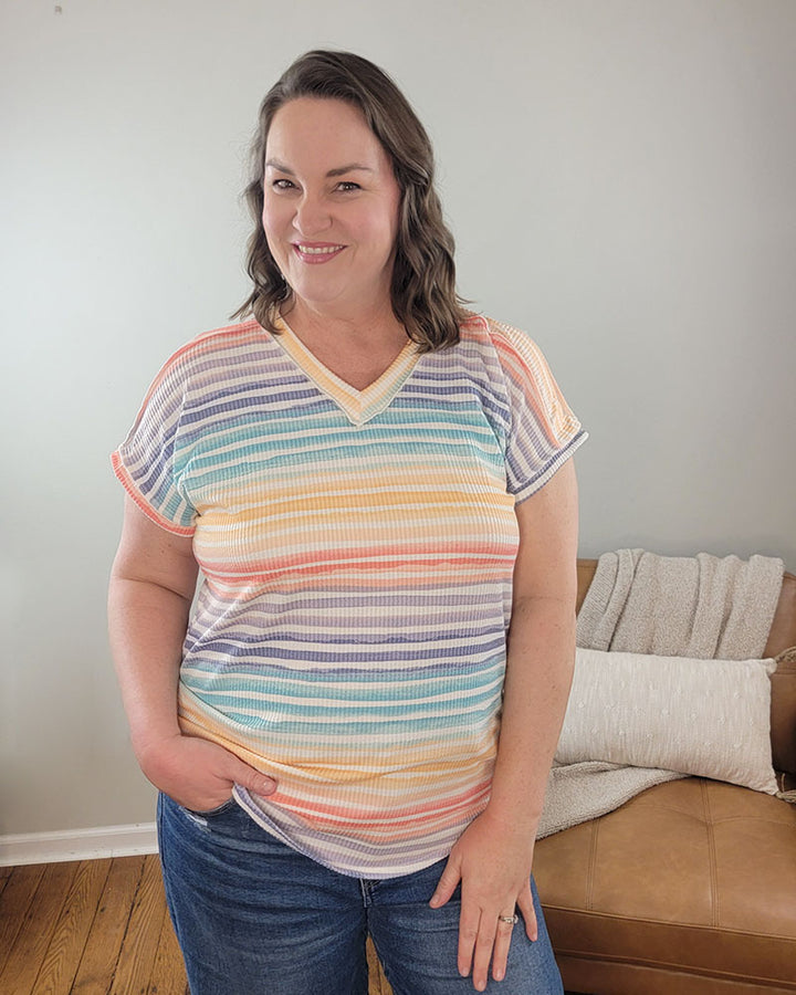 A woman with wavy brown hair smiles at the camera indoors, wearing the Heimish Hilary Striped V-Neck and blue jeans. One hand is in her pocket, and a tan couch with a blanket appears in the background.
