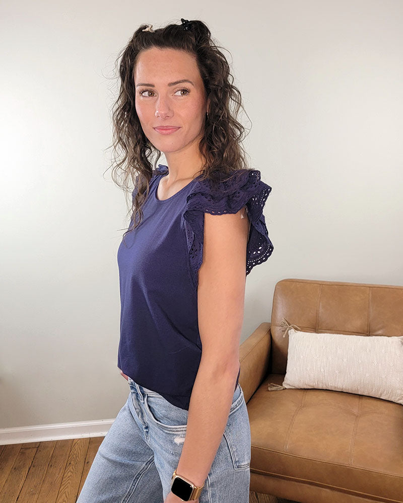 A woman with wavy brown hair wears the Staccato Preston Top, a navy blue blouse with flutter sleeves, paired with light blue jeans. She stands indoors by a brown sofa with a white pillow, gazing slightly to the side.