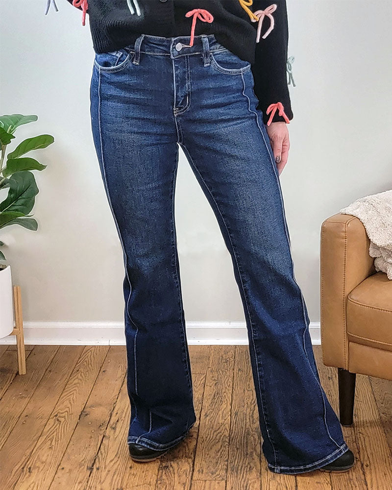 A person wears Lovervet’s Chicness Flare Jeans and a black sweater with colorful bows, standing indoors on a wooden floor near a green plant and brown chair.