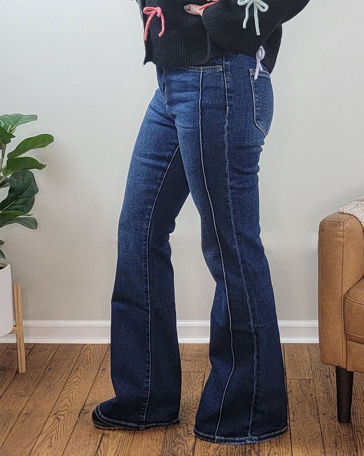 A person stands indoors wearing Lovervet’s Chicness Flare Jeans in dark wash comfort stretch denim and a black sweater with pastel ribbon sleeve details. A plant in a white pot and part of a tan chair appear in the background.