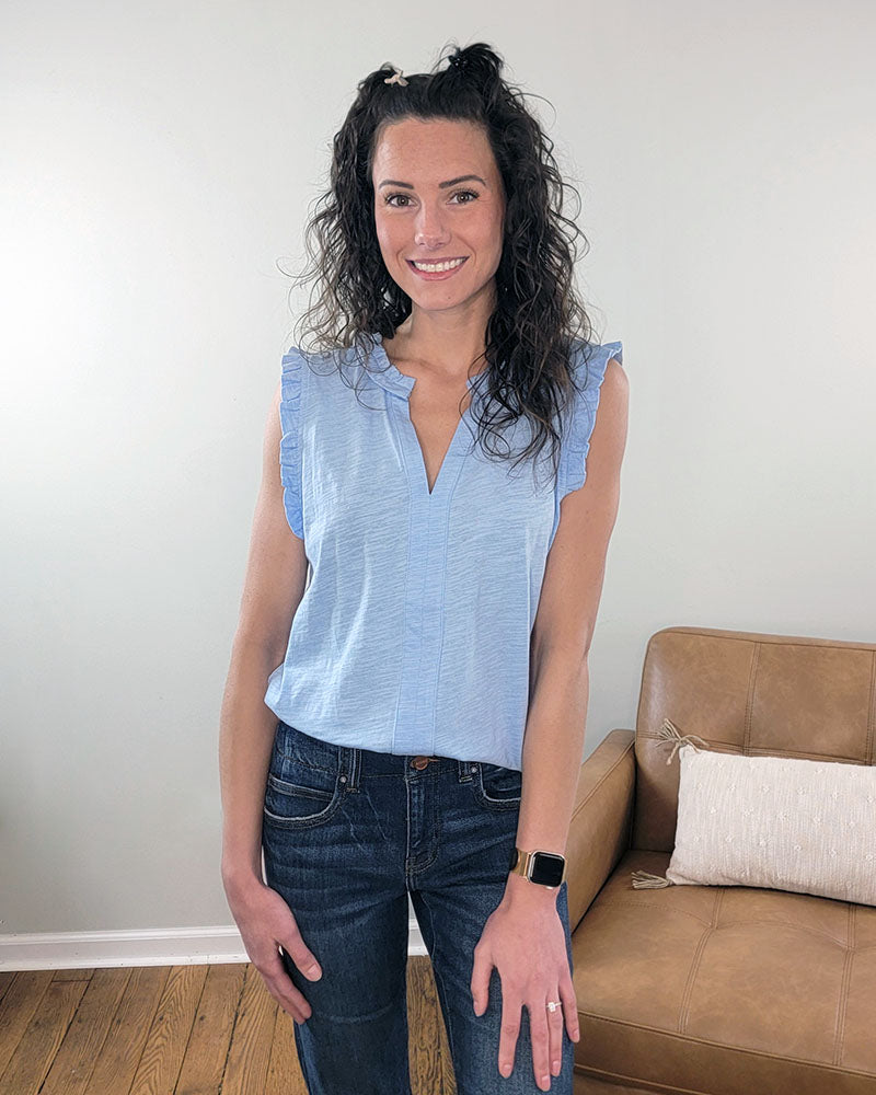 A woman with curly dark hair smiles in a living room, wearing the Staccato Illa V-Neck Sleeveless Top with Ruffle Details and jeans. A tan couch and cushion are visible in the background.
