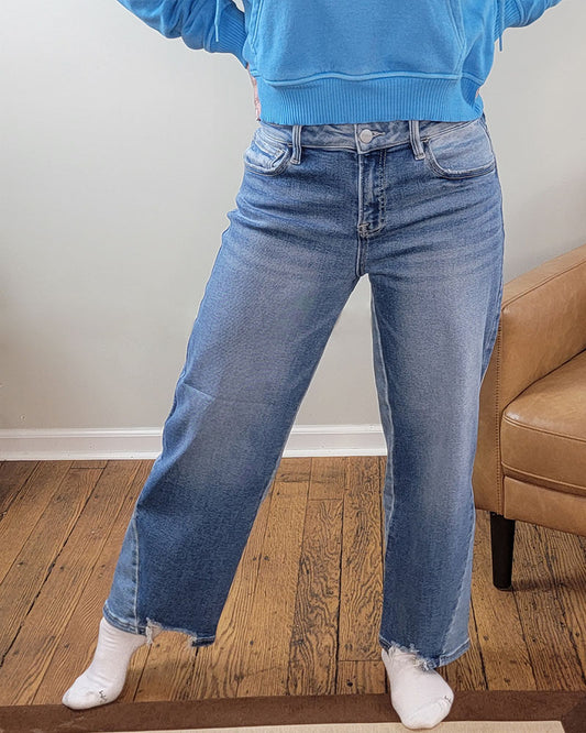 A person stands indoors on a wooden floor, one leg slightly forward, wearing Risen's Astrid Slouchy Crop Barrel Jeans with a raw hem, a blue sweatshirt, and white socks. A brown chair is visible to the side.