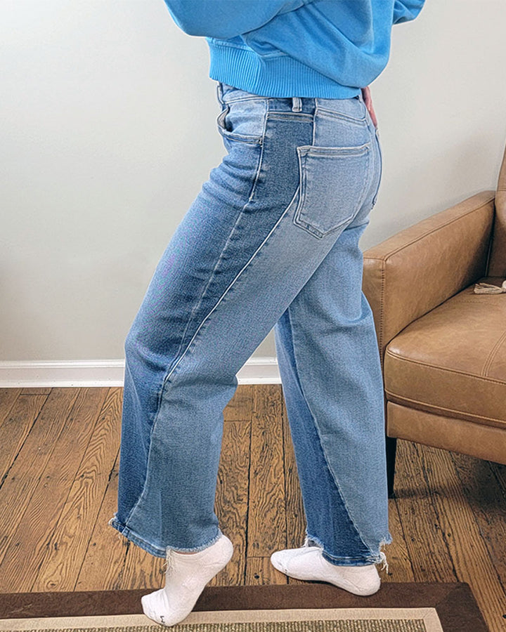 Someone stands on a wooden floor by a brown armchair, wearing Risen Astrid Slouchy Crop Barrel Jeans in light blue with frayed hems and a blue sweatshirt, showing the side and back views while wearing white socks.