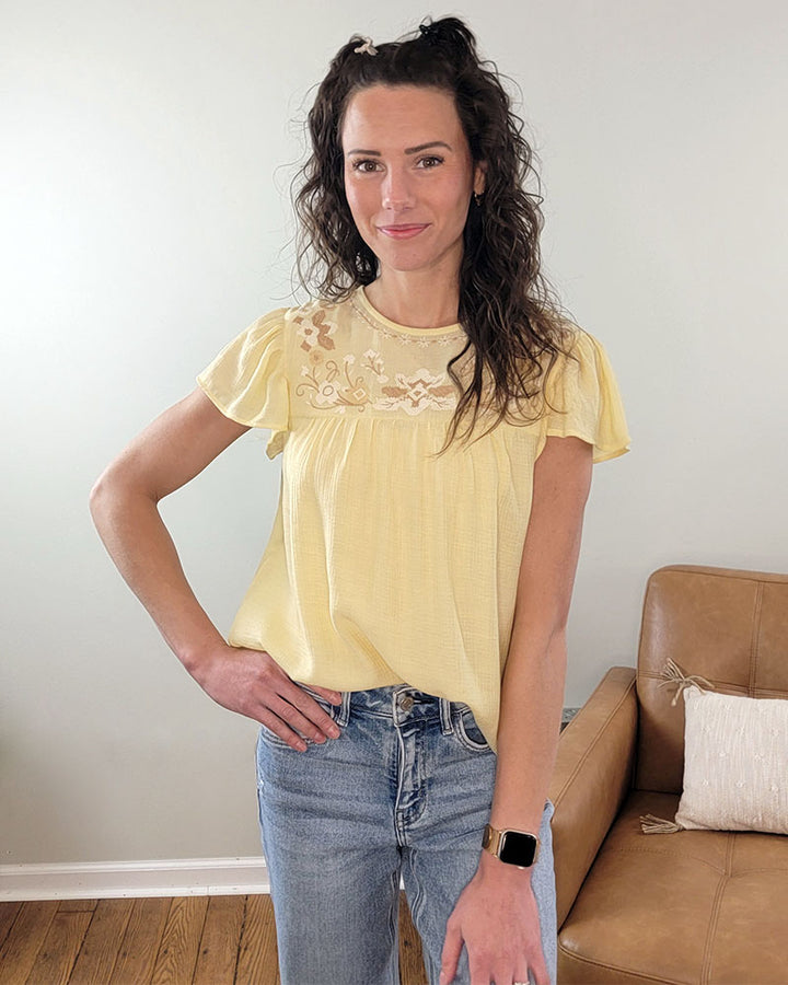 A woman with long curly hair smiles indoors, wearing the Mine and E&M Addison Flutter Sleeve Top in butter yellow with embroidery and blue jeans. A brown chair with a pillow sits behind her.