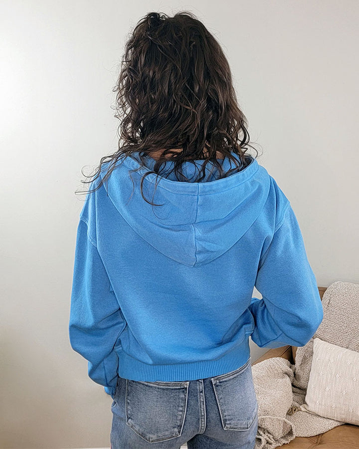Wearing the ZENANA Maeve Half Zip Cropped Hoodie, a person with long, wavy brown hair and light blue jeans stands with their back to the camera in a minimal, light-colored room.