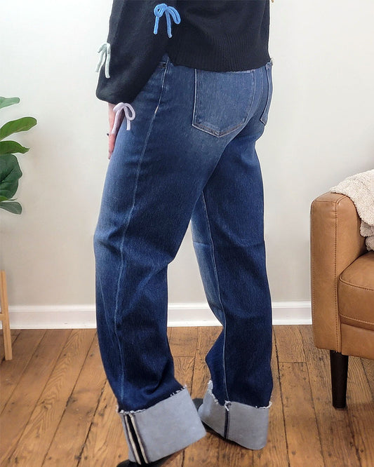 Wearing Meadow Wide Straight Jeans from Risen with rolled cuffs and a black sweater featuring blue and purple bows, the person stands on a wooden floor near a tan chair and green plant, highlighting stylish denim by Risen.