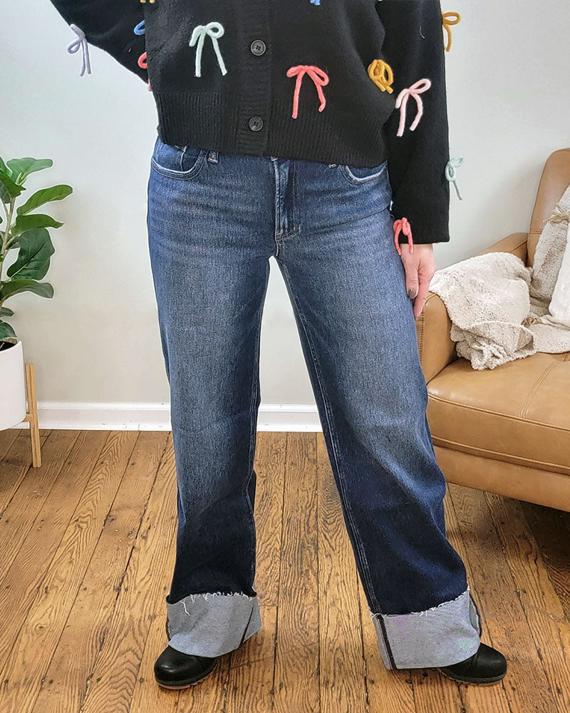 Indoors on a wooden floor, a person wears Risen's Meadow Wide Straight Jeans with large cuffs, black shoes, and a black sweater featuring colorful bows. A plant and chair are in the background.