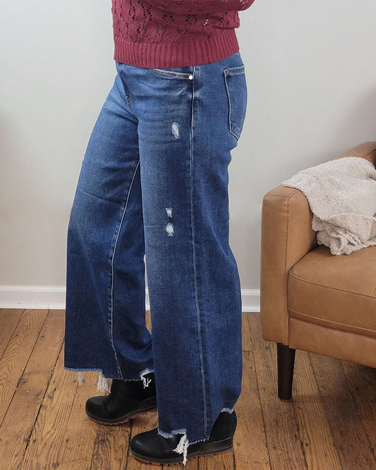 A person in a red sweater and Risen's Briar Crop Wide Leg Jeans, featuring frayed hems and distressed details, stands on a wooden floor beside a tan armchair.
