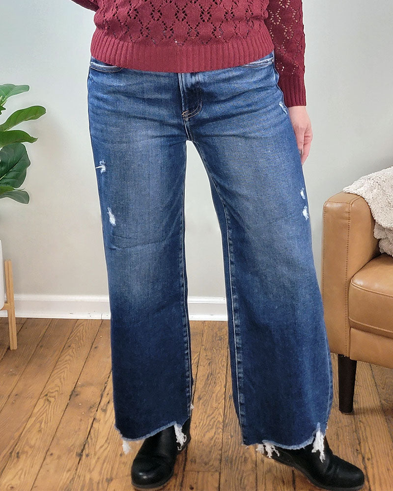 A person wears maroon knit sweater and Briar Crop Wide Leg Jeans by Risen, standing indoors on a wooden floor near a tan chair and green plant.