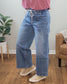 A person, only visible from the waist down, stands on a wooden floor beside a potted plant and tan armchair, modeling stylish high rise Reassure Wide Leg Jeans from Vervet with a drawstring waist and paired with a pink top.