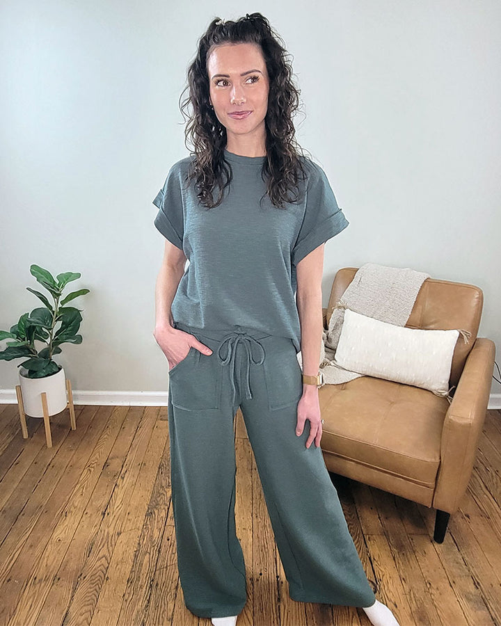 A woman with curly hair stands indoors wearing the ZENANA Daphne Top and Pant Set in green, featuring short sleeves and elastic waist pants with pockets. Behind her are a tan cushioned chair and a potted plant on a wooden floor.