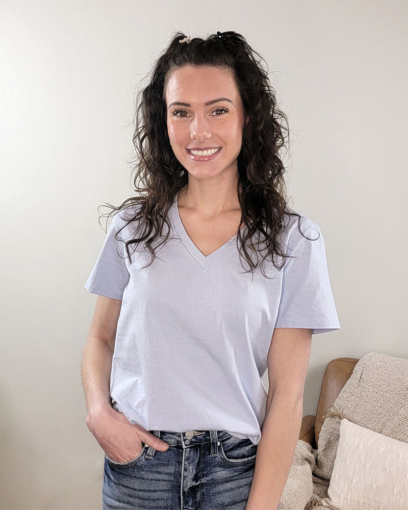 A woman with long, curly brown hair smiles indoors, wearing the Yelete Group Emilia V-neck Tee in light purple and blue jeans. A beige pillow and textured blanket are on the couch behind her.