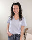 A woman with long, curly brown hair smiles indoors, wearing the Yelete Group Emilia V-neck Tee in light purple and blue jeans. A beige pillow and textured blanket are on the couch behind her.