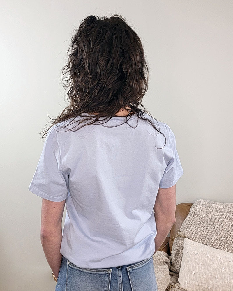 A person with long, wavy brown hair stands indoors facing away, wearing the Emilia V-neck Tee by Yelete Group in light blue and light blue jeans. Behind them is a beige couch with textured cushions.