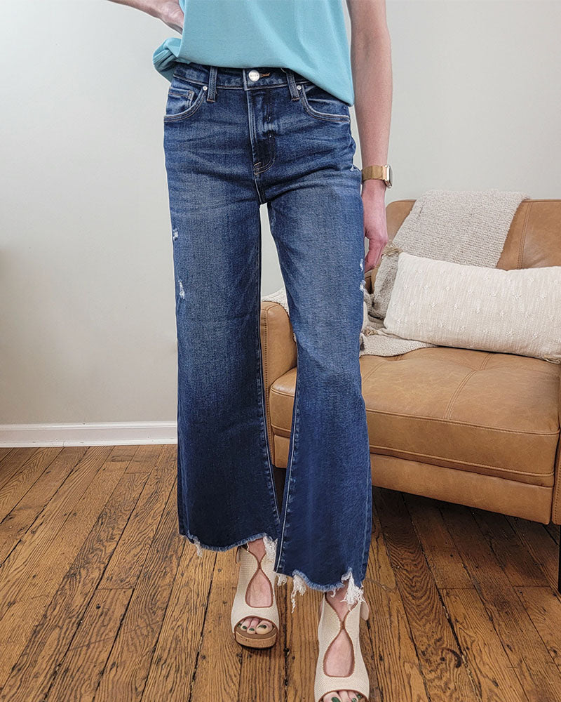 A person wearing a light blue sleeveless top, Briar Crop Wide Leg Jeans by Risen, and beige heeled sandals stands on a wooden floor beside a tan couch with pillows.