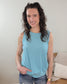 A woman with curly brown hair, wearing the powder blue Ayla Ribbed Tank by Cosmos + Clover Boutique and jeans, stands indoors smiling at the camera with one hand in her pocket. A beige couch with pillows is visible in the background.