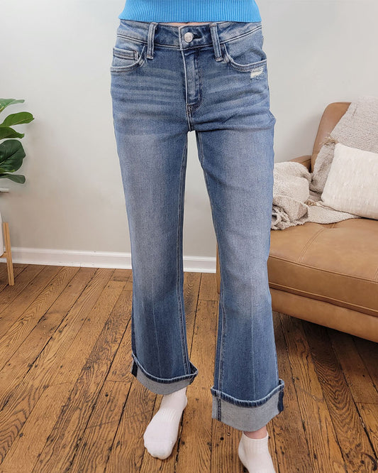 A person wearing Lovervet’s Delicate Gleam Cuffed Slim Wide Leg Jeans stands on a wooden floor near a tan couch with beige accents; a green plant appears in the background.