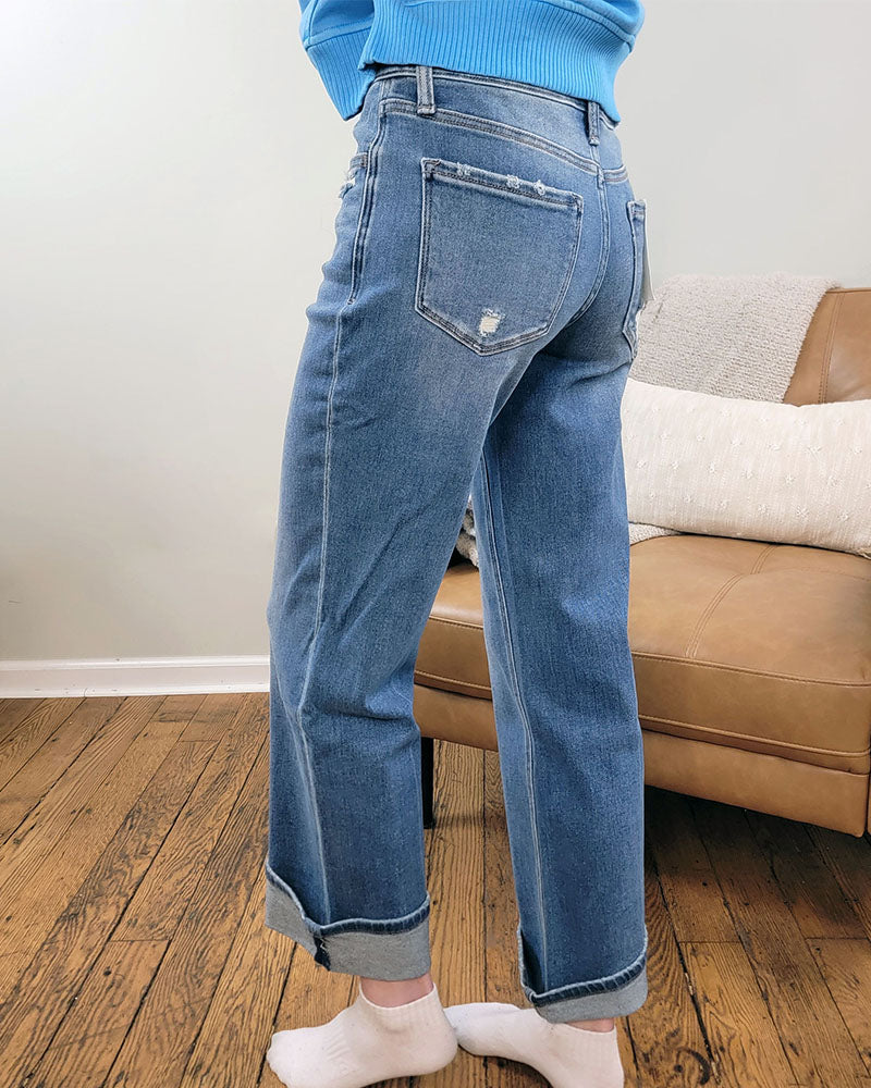 A person wearing Lovervet's Delicate Gleam Cuffed Slim Wide Leg Jeans stands on a wooden floor near a tan couch with white pillows, facing away from the camera.