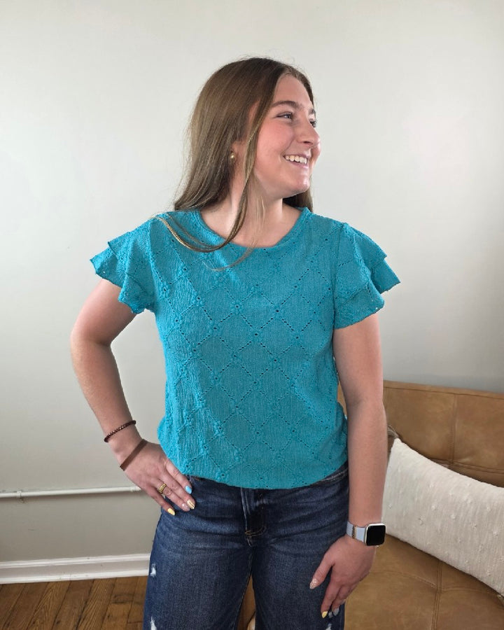 A woman with long brown hair stands indoors, smiling to the side with one hand on her hip. She wears blue jeans and the Lovesoft Wren Eyelet &amp; Ruffle Sleeve Top in bright turquoise, along with an Apple Watch on her wrist.