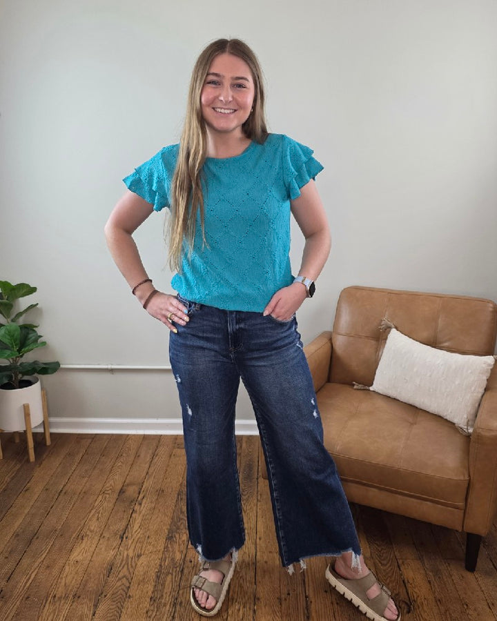 A woman with long blonde hair, wearing the Lovesoft Wren Eyelet &amp; Ruffle Sleeve Top, wide-leg dark jeans, and sandals, smiles in a living room featuring wood floors, a tan armchair, and a potted plant.