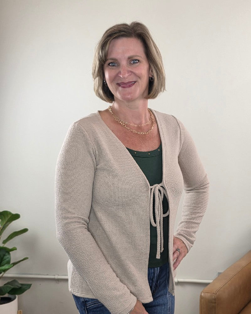 A woman with short blonde hair smiles at the camera indoors, wearing the Les Amis Wrenlee Tie Front Knit Cardigan in taupe over a green top and jeans. A potted plant and part of a brown couch are visible in the background.