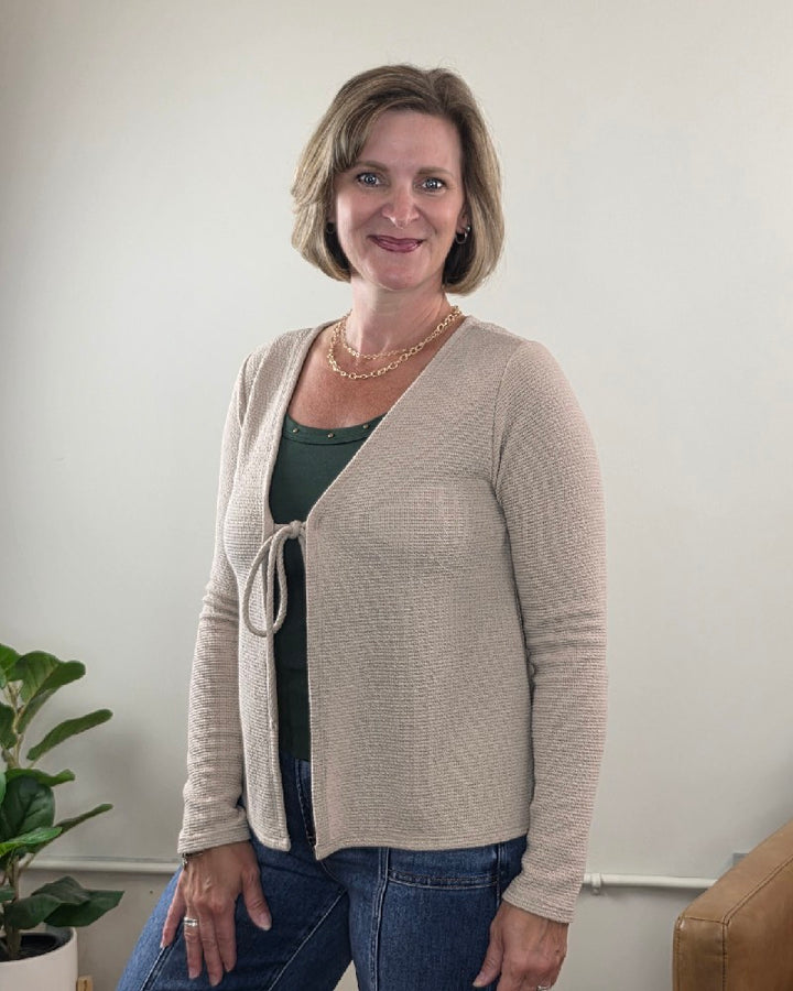A woman with short brown hair, wearing the Les Amis Wrenlee Tie Front Knit Cardigan in taupe over a green top and blue jeans, stands indoors near a plant and a tan couch, smiling at the camera.