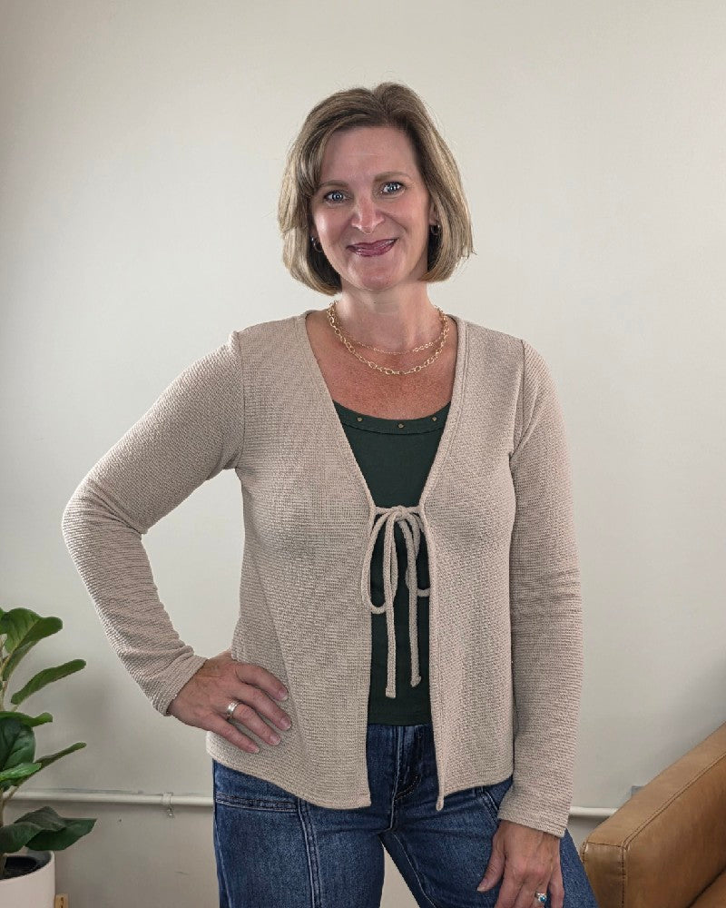 A woman with short blonde hair is smiling indoors, standing with her hand on her hip. She’s wearing the Les Amis Wrenlee Tie Front Knit Cardigan in taupe over a dark green shirt. In the background, there’s a plant and a tan couch.