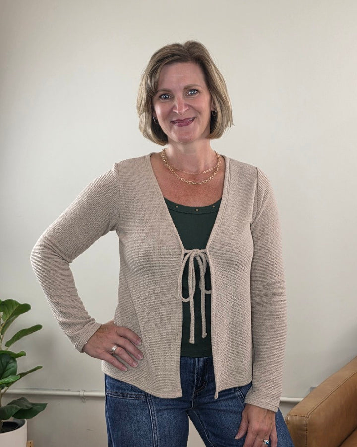 A woman with short blonde hair is smiling indoors, standing with her hand on her hip. She’s wearing the Les Amis Wrenlee Tie Front Knit Cardigan in taupe over a dark green shirt. In the background, there’s a plant and a tan couch.