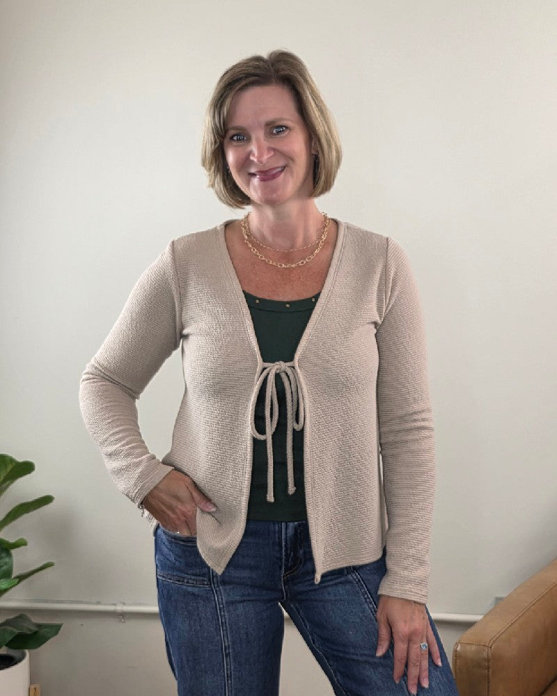 A woman with short blonde hair smiles while standing indoors, wearing the Les Amis Wrenlee Tie Front Knit Cardigan in taupe over a green top and blue jeans. She has one hand on her hip and stands next to a plant and a tan couch.
