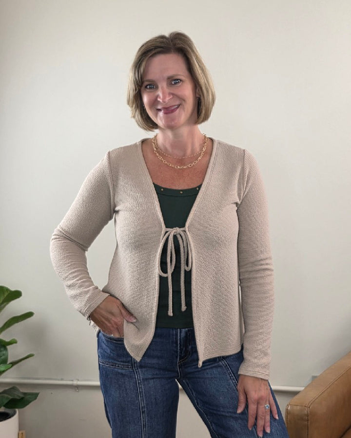 A woman with short blonde hair smiles while standing indoors, wearing the Les Amis Wrenlee Tie Front Knit Cardigan in taupe over a green top and blue jeans. She has one hand on her hip and stands next to a plant and a tan couch.