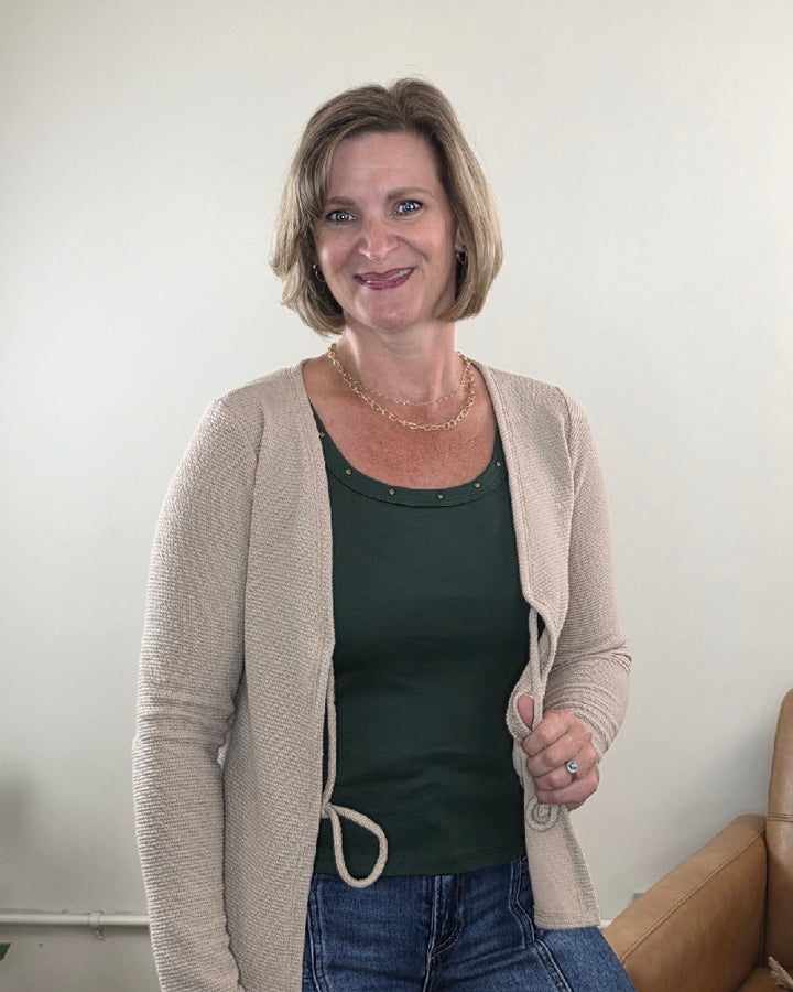 A woman with short light brown hair wears the Timing Jessica Studded Tank Top in dark green, layered under a beige cardigan and paired with blue jeans. She stands smiling in a well-lit room, with a tan chair visible in the background.