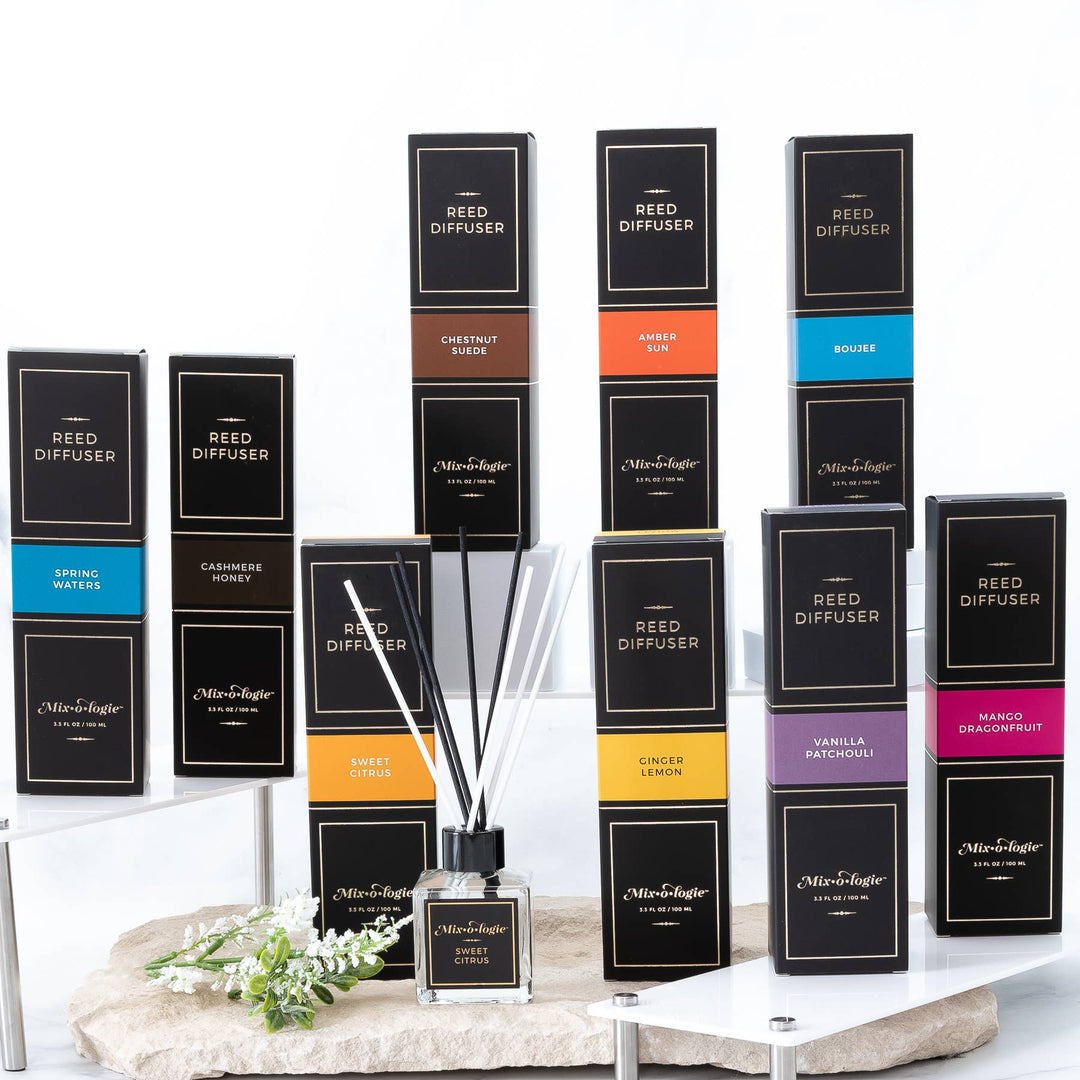 A display of twelve Mixologie Reed Diffusers boxes in assorted colors and scents is tiered with a diffuser bottle and sticks in front—ideal as chic hostess gifts—all featured against a white background.