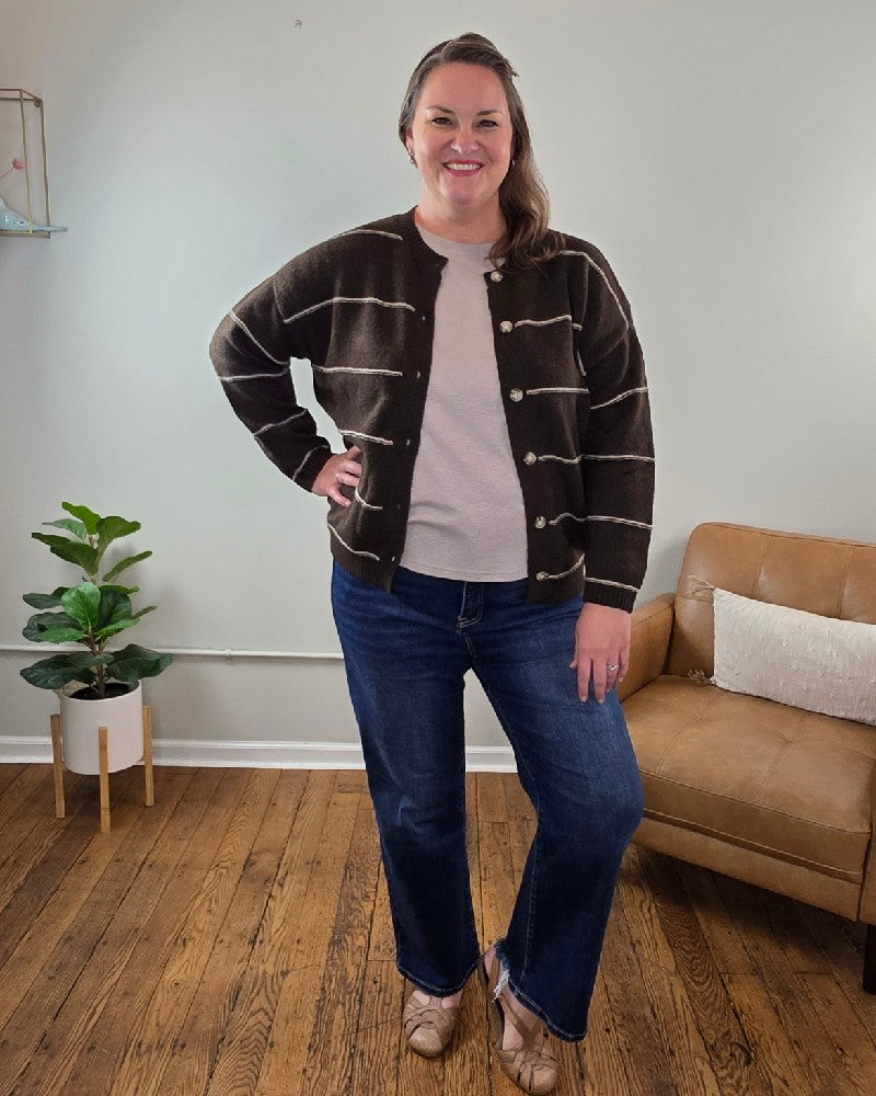 A woman stands indoors on a wooden floor, wearing the blu pepper Aliza Striped Button Down Sweater with blue jeans and tan shoes. She smiles with one hand on her hip; a small plant and brown chair are in the background.