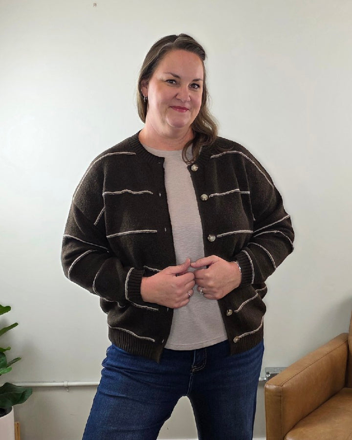 A woman with long brown hair smiles indoors, wearing the blu pepper Aliza Striped Button Down Sweater over a light shirt and jeans, lightly holding its edges.