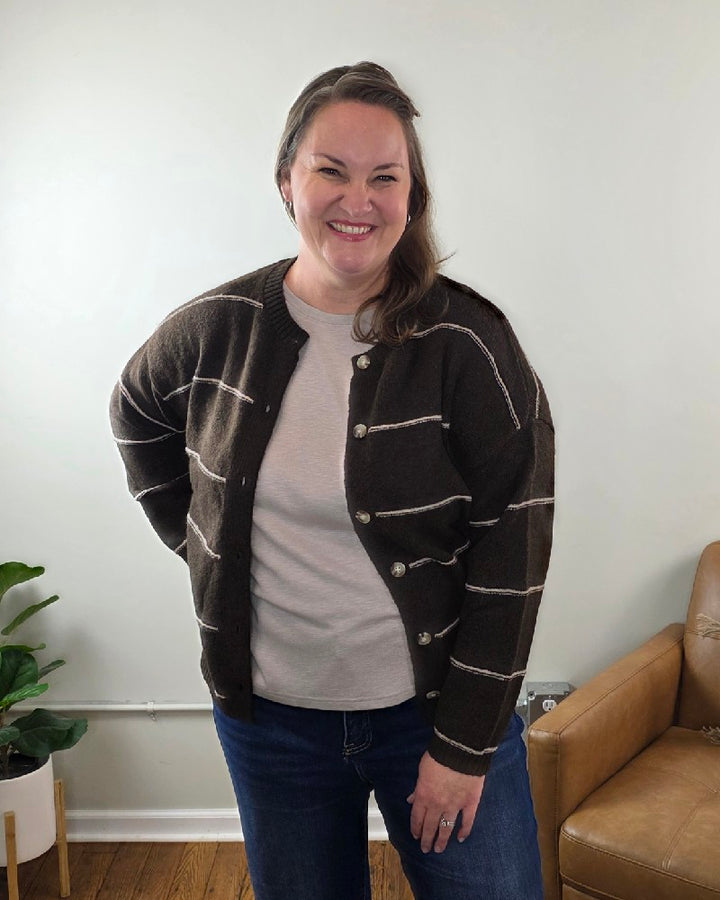 A woman with long brown hair, wearing the blu pepper Aliza Striped Button Down Sweater and blue jeans, smiles in a living room with a leather chair, a plant, and light-colored walls.