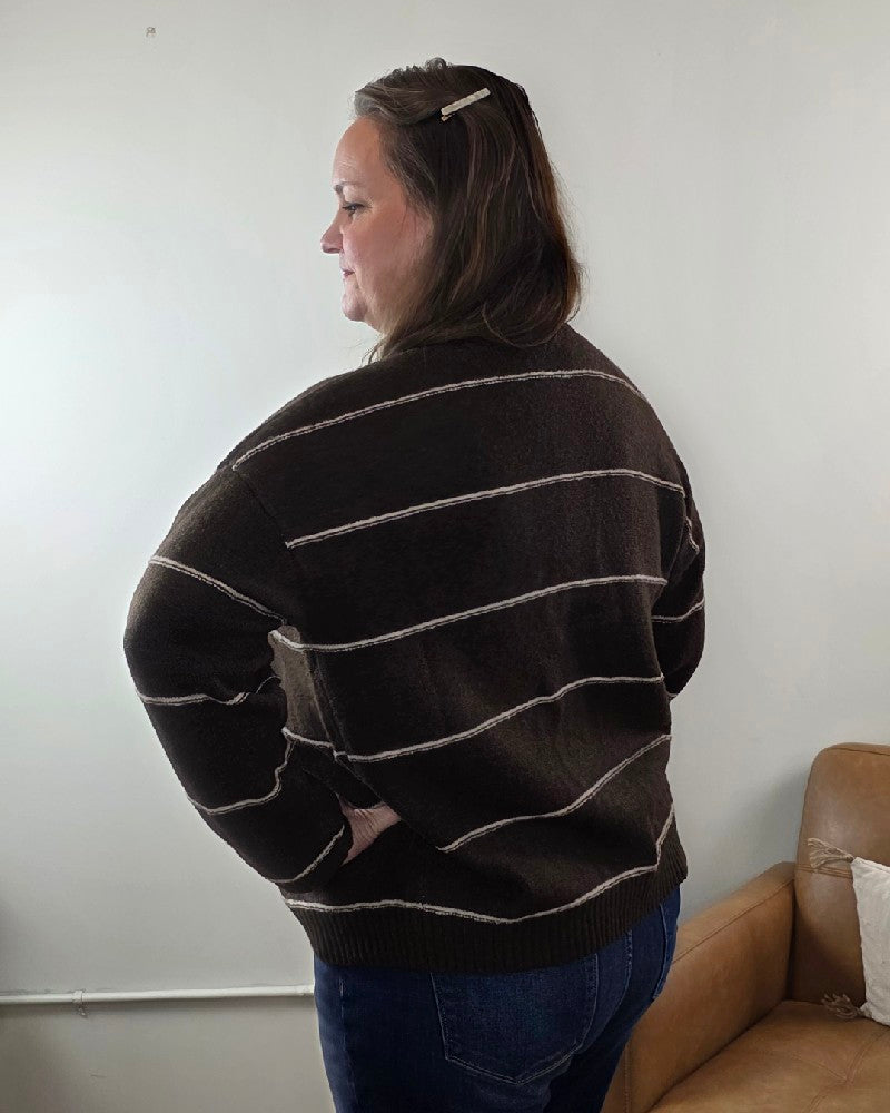A woman with brown hair clipped back stands sideways, wearing the Aliza Striped Button Down Sweater by blu pepper with blue jeans, indoors near a light wall and tan couch.