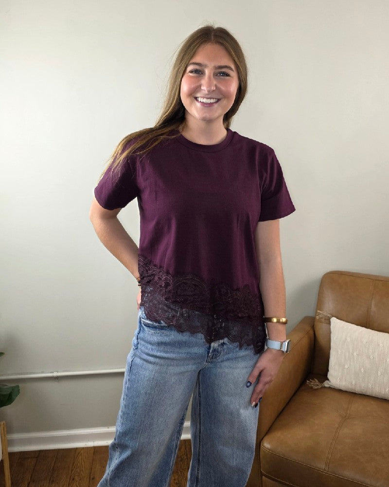 A woman wearing an Amiri Lace Edge Top in dark purple with a lace hem and blue jeans.