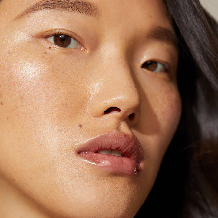 Close-up of a woman with smooth, glowing skin, natural makeup, and long black hair. She wears Minori Lip Gloss - Cozy in a bronzy nude shade, highlighting her light freckles and mole as she embraces clean beauty with a calm expression.
