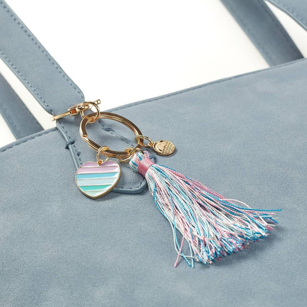 A close-up of the Carrie Keychain by OOLY, a blue suede handbag accessory with a gold keyring and lobster clasp, featuring a pastel striped heart enamel charm, gold circular charm, and blue and pink tassel.