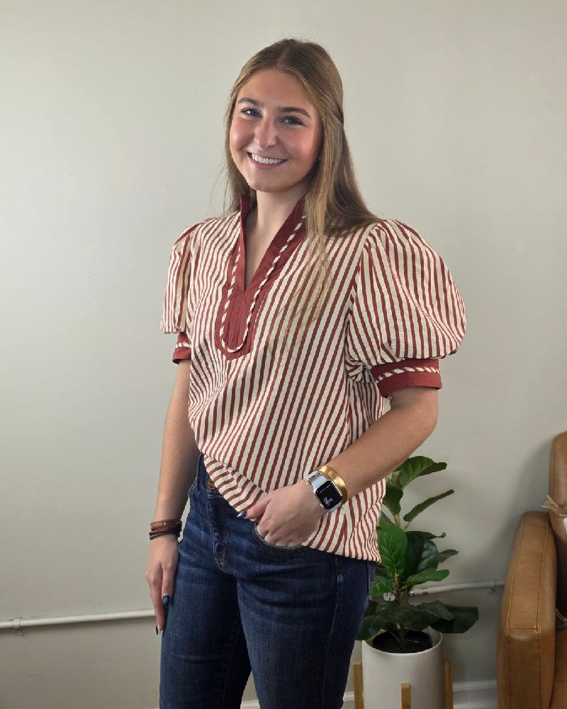A smiling woman wears a red and white vertical stripe Brittany V neck Puff Sleeve Stripe Blouse with puff sleeves and blue jeans.