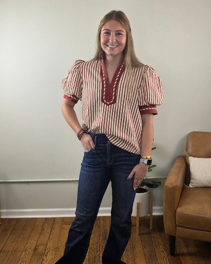 A woman wearing a rust striped Brittany V neck Puff Sleeve Stripe Blouse with a V-neck and short puff sleeves, paired with dark wash jeans.