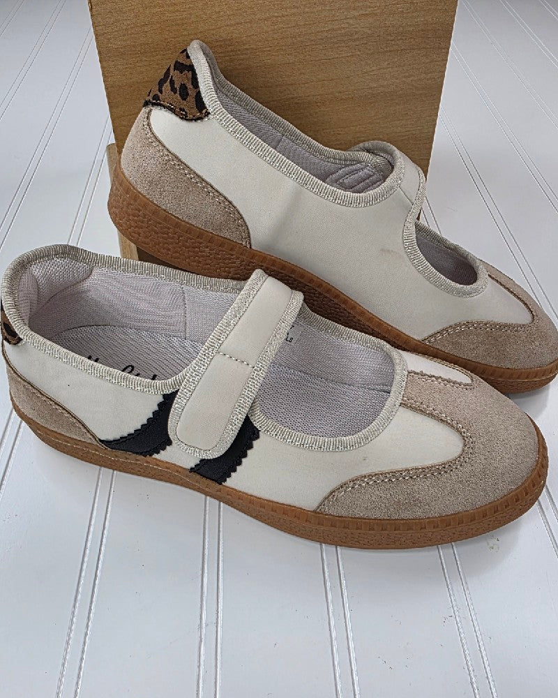 The Calm Down Sand Combo Slip on Shoe by Corkys features a mix of beige and tan in a classic Mary Jane silhouette, accented with black and beige details, a convenient Velcro strap, brown rubber soles, and a subtle leopard print accent on the heel. Presented on a white surface with a wooden backdrop, this stylish neutral sneaker offers the perfect blend of comfort and versatility for an effortlessly chic look.